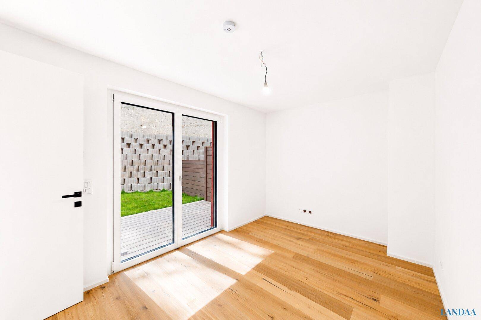 Heller, leerer Raum mit Holzboden und großen Glasfronten zum Garten.