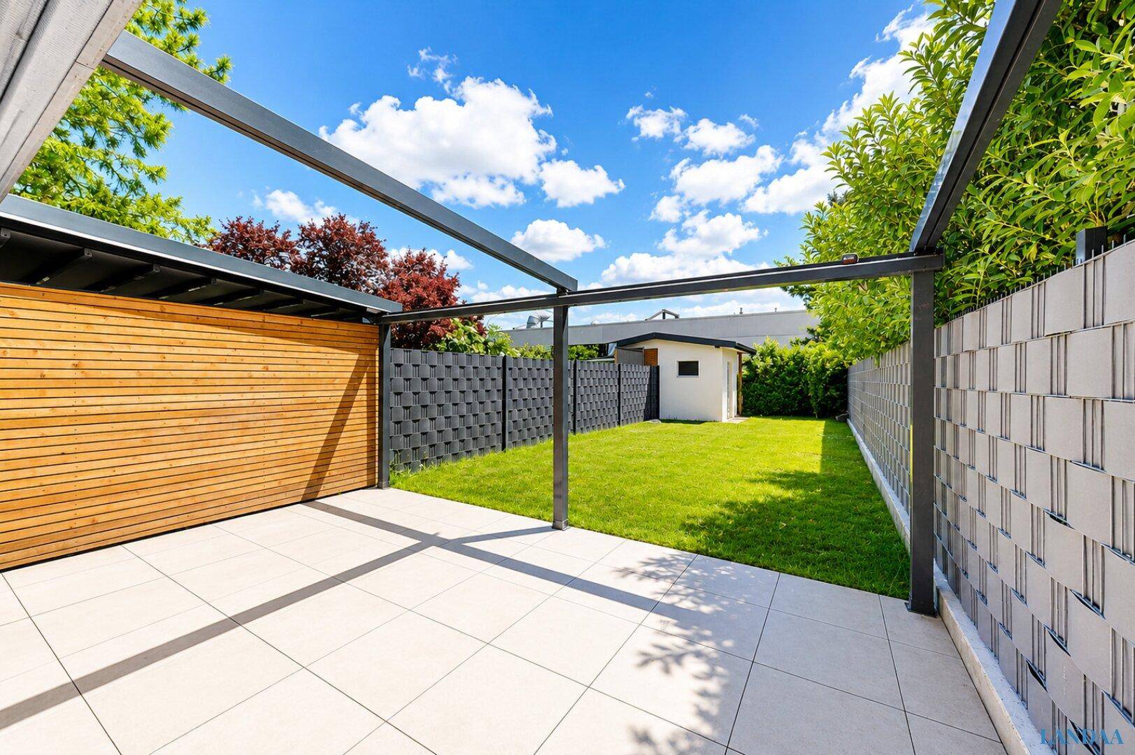 Sonniger Terrassenbereich mit Fliesenboden, Pergola und Blick auf den kleinen Garten.