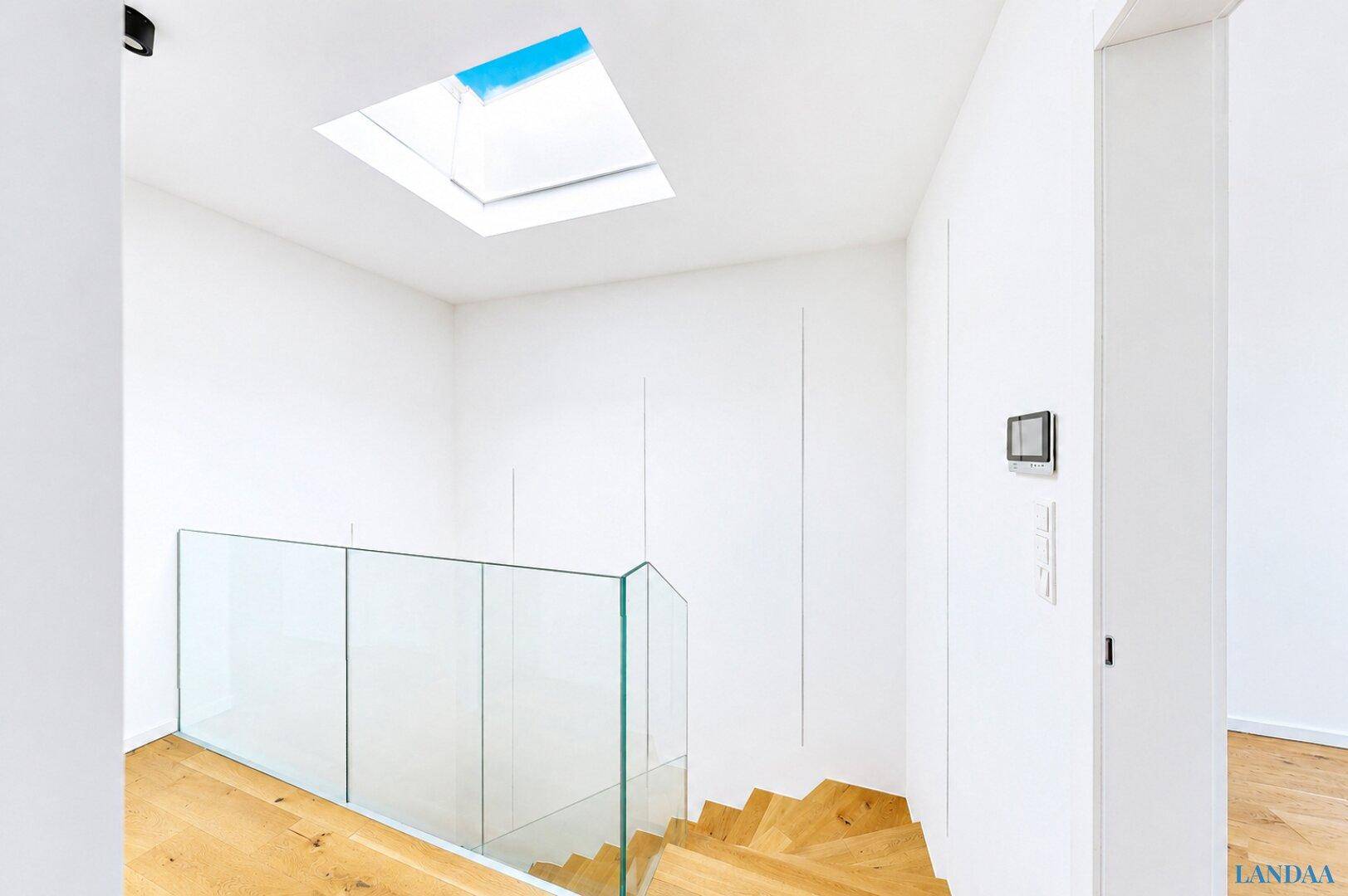 Heller Treppenbereich mit Oberlicht, Glasgeländer und weiß gestrichenen Wänden.