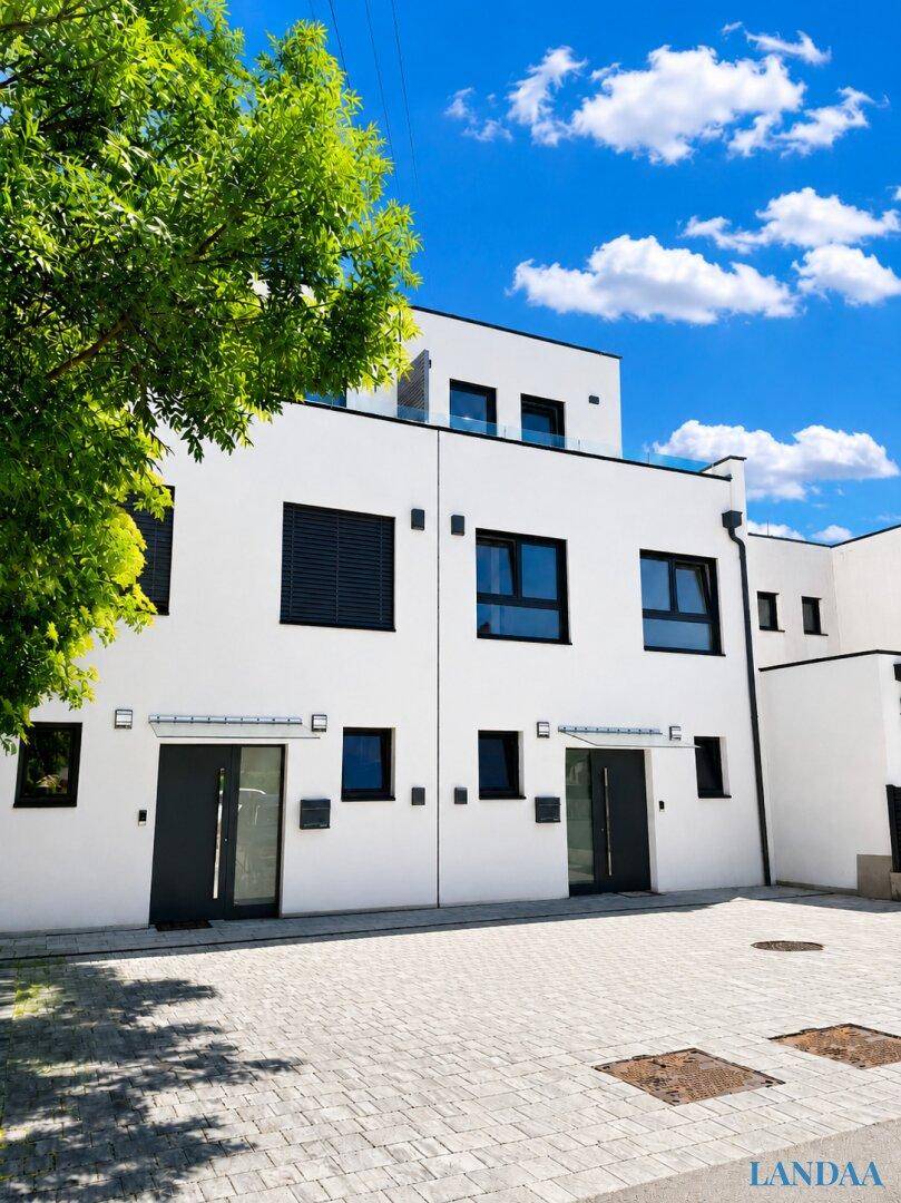Moderne Reihenhausfassade mit großen Fenstern und gepflastertem Vorplatz unter blauem Himmel.