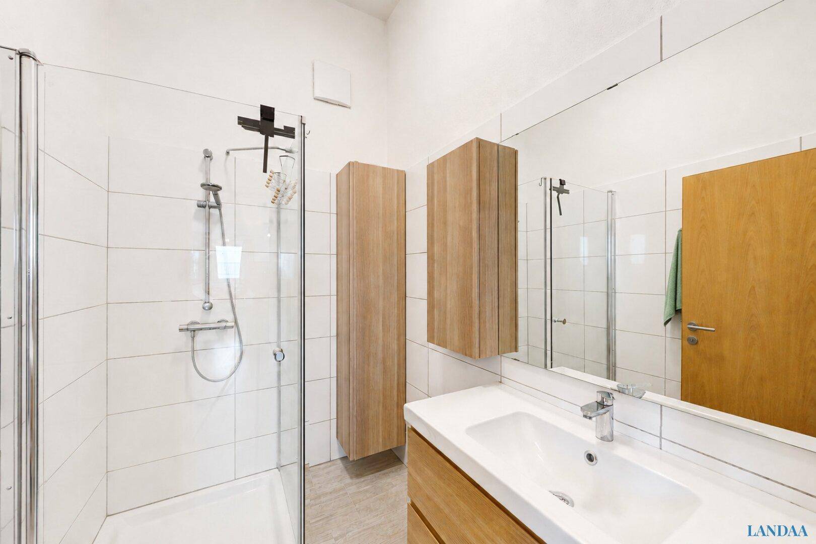 Modernes Badezimmer mit Glasduschkabine, Waschtisch, großem Spiegel und Holzschränken an weißen Fliesenwänden.