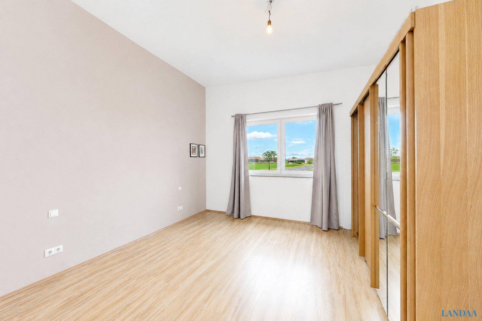 Leeres Schlafzimmer mit hellem Laminatboden, beigen Wänden, Fenster mit Vorhängen und großem Spiegelschrank.