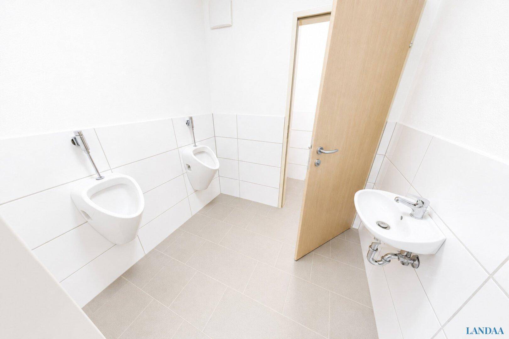 Herrentoilette mit zwei Urinalen, kleinem Waschbecken, weißen Fliesenwänden und einer Holztür.