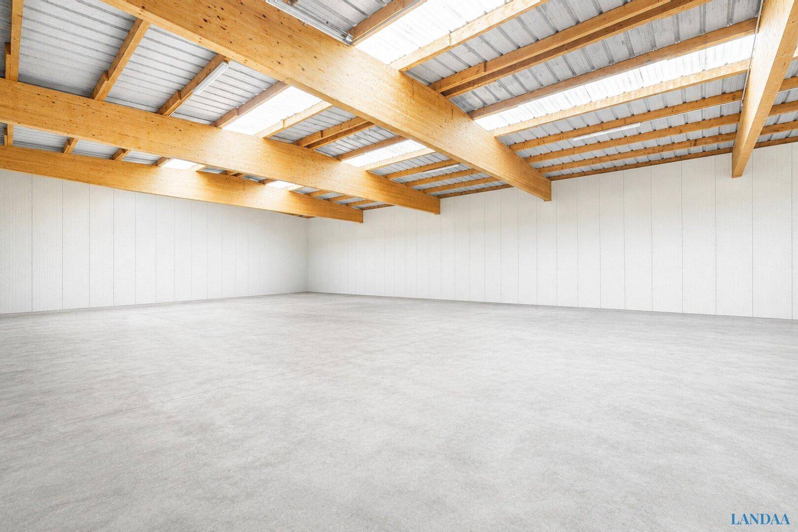 Weitläufige, leere Lagerhalle mit hohen Decken, Holzbalken und viel natürlichem Licht durch die Oberlichter.