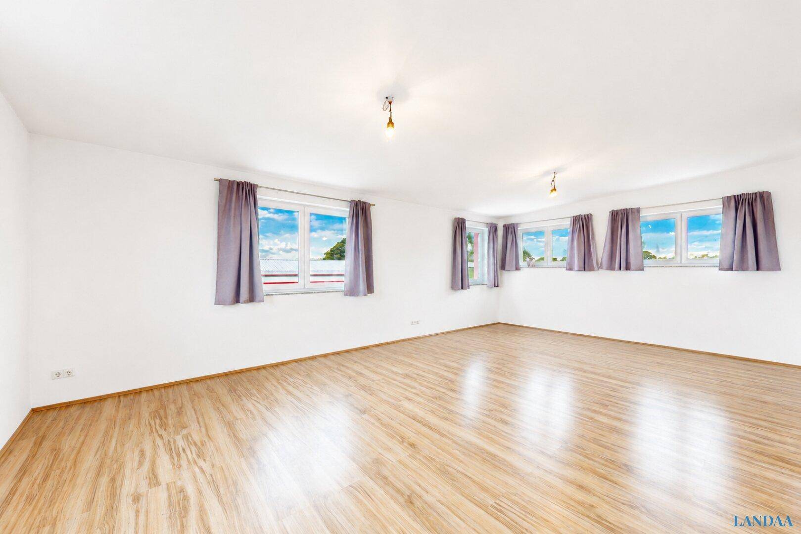 Geräumiges Wohnzimmer mit hellem Laminatboden, weißen Wänden und mehreren Fenstern mit Vorhängen.