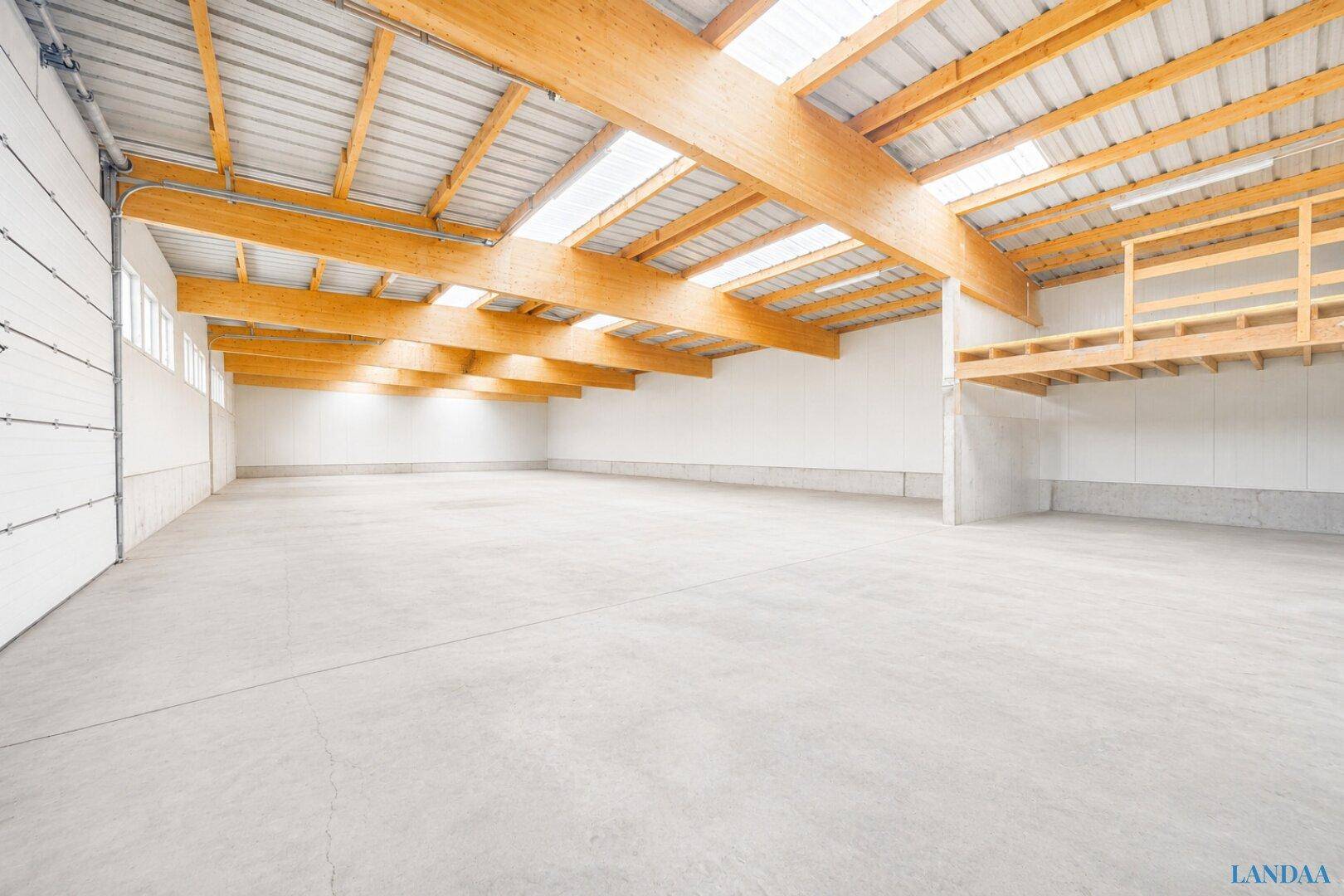 Geräumige, leere Lagerhalle mit hohen Decken, Holzbalken, Oberlichtern und einer sichtbaren Zwischenebene.
