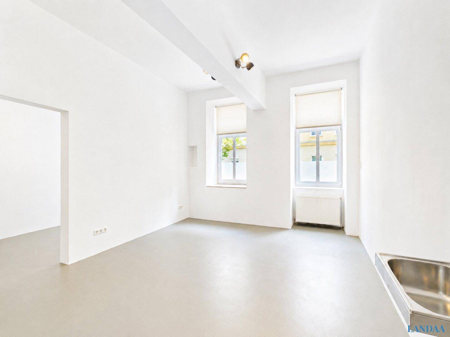 Heller, leerer Raum mit zwei Fenstern, hellem Boden und einem kleinen Waschbecken in der Ecke.