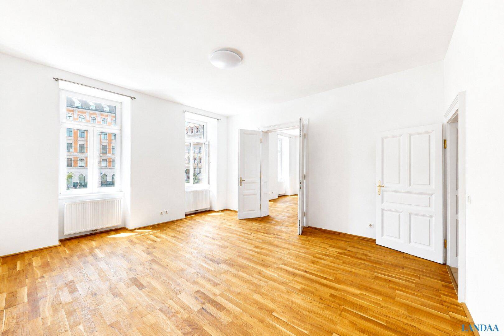 Lichtdurchfluteter Raum mit Fischgrätparkett und hohen Türen, der mehrere Zimmer verbindet.