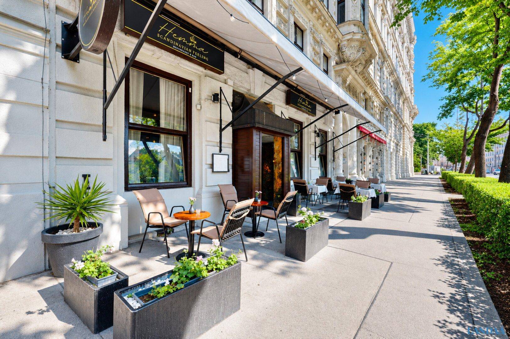 Breite Ansicht der Restaurantterrasse mit Tischen, Stühlen und Pflanzenkübeln entlang des Bürgersteigs.