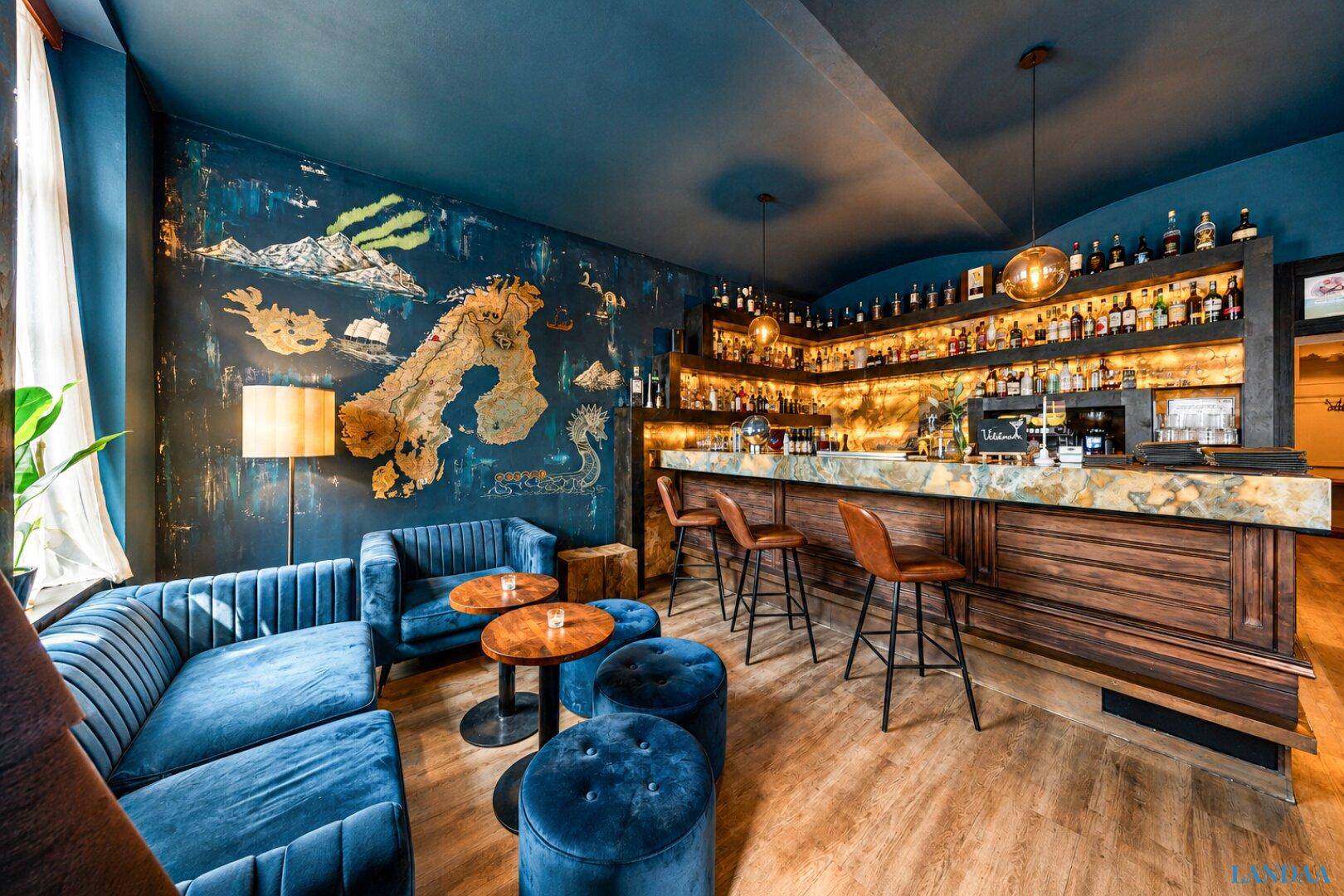 Gemütliche Bar mit blauen Samtsofas, Holzboden und einer reich bestückten Theke mit Flaschen.