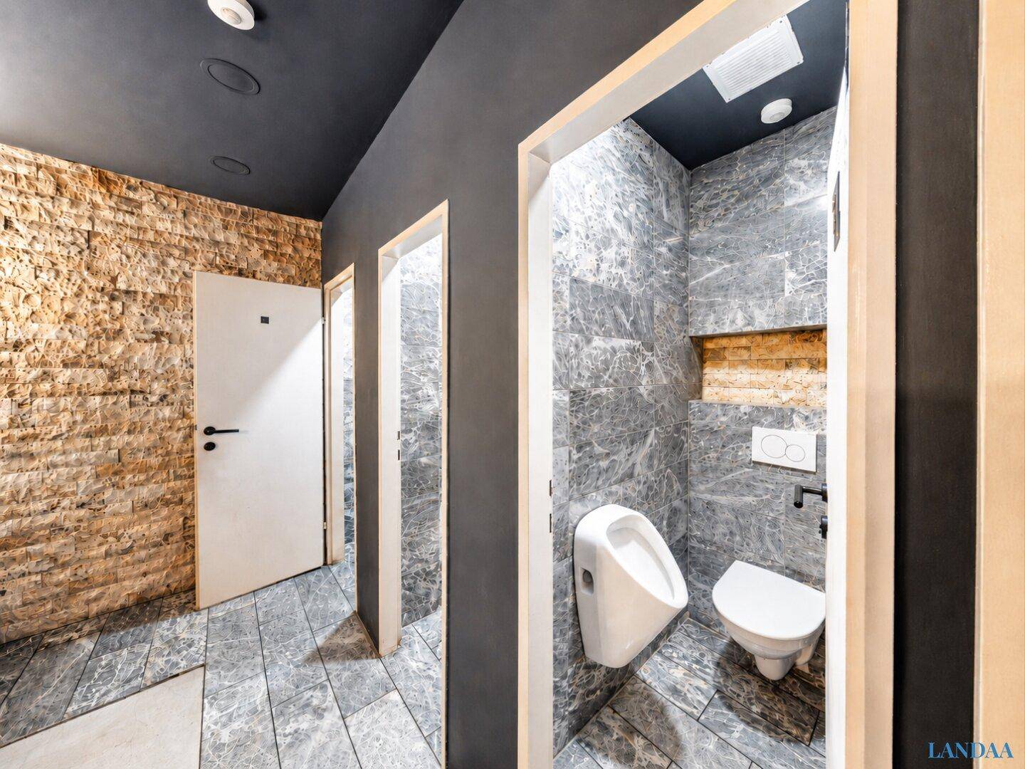 Toilettenbereich mit Urinal und WC, grauen Marmorfliesen und einer strukturierten Wand.