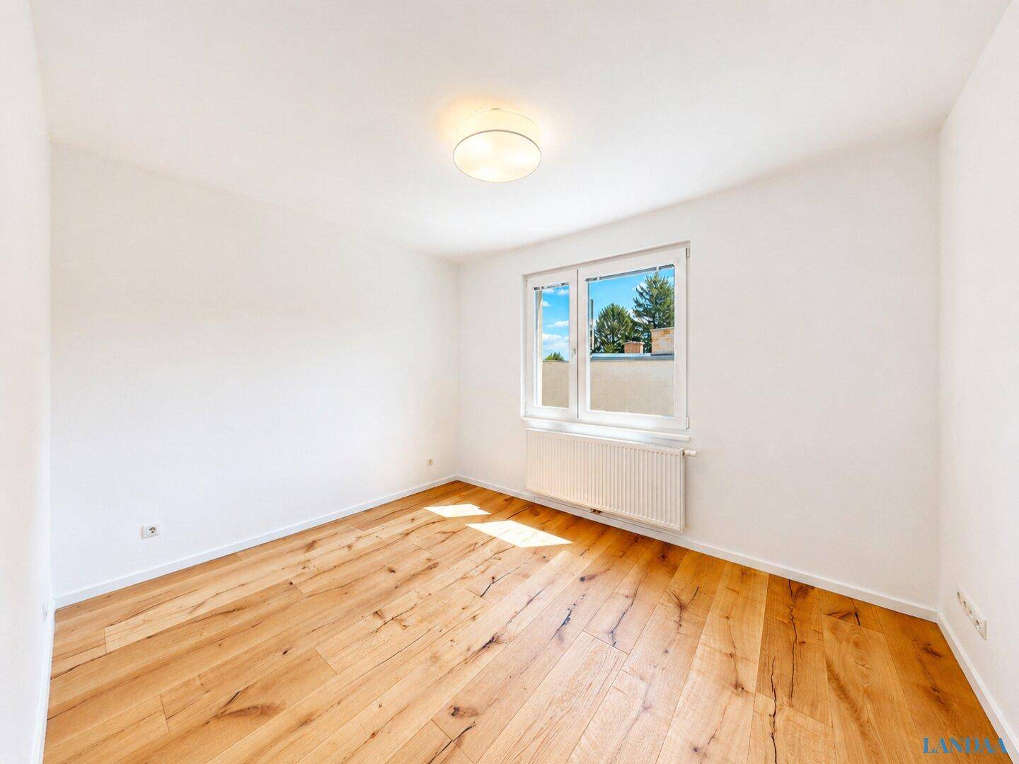 Helles, leeres Schlafzimmer mit Holzfußboden und einem Fenster, das viel Tageslicht hereinlässt.