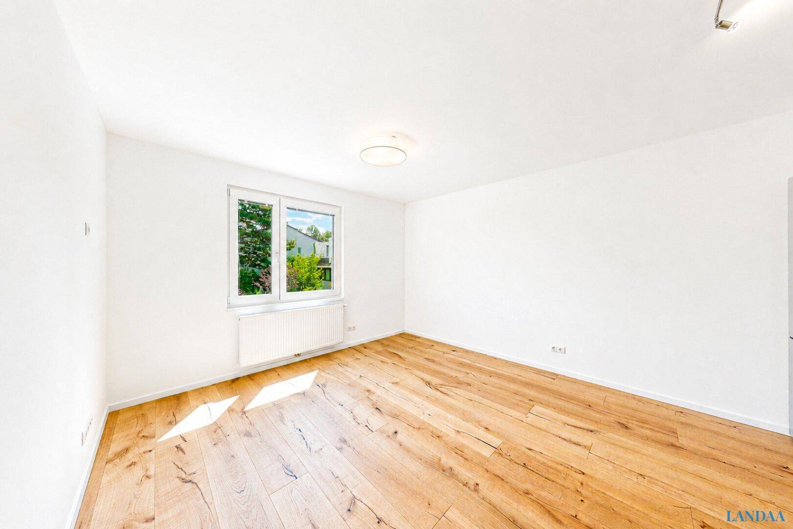 Leeres Zimmer mit hellem Holzfußboden und einem Fenster mit Blick auf grüne Bäume.