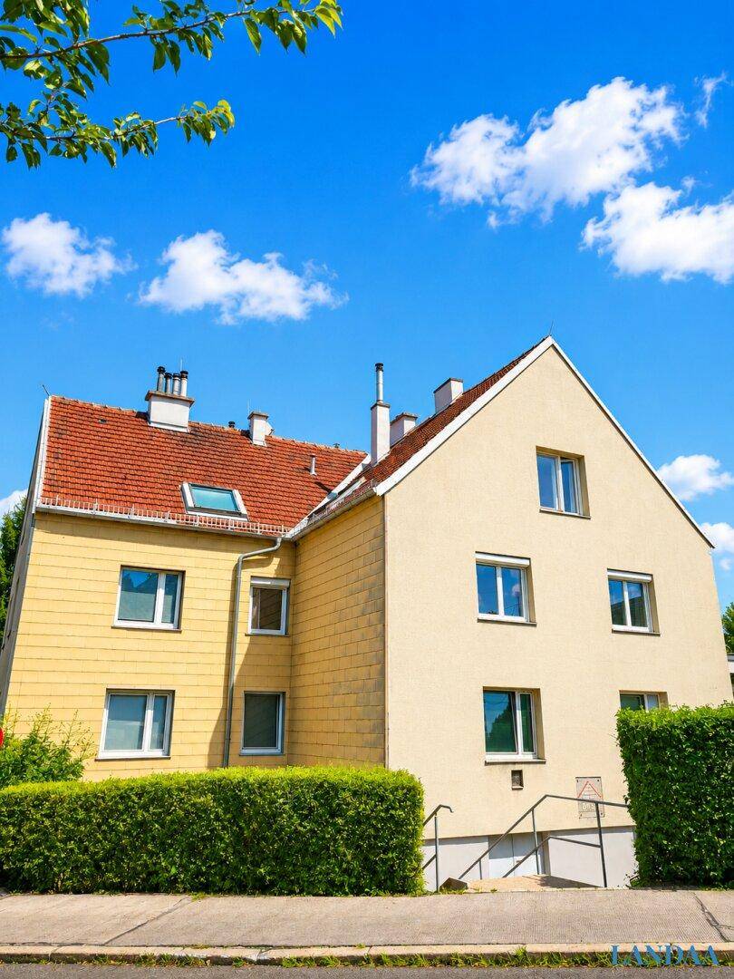 Ansicht eines Mehrfamilienhauses mit gelber Fassade, rotem Ziegeldach und grüner Hecke.