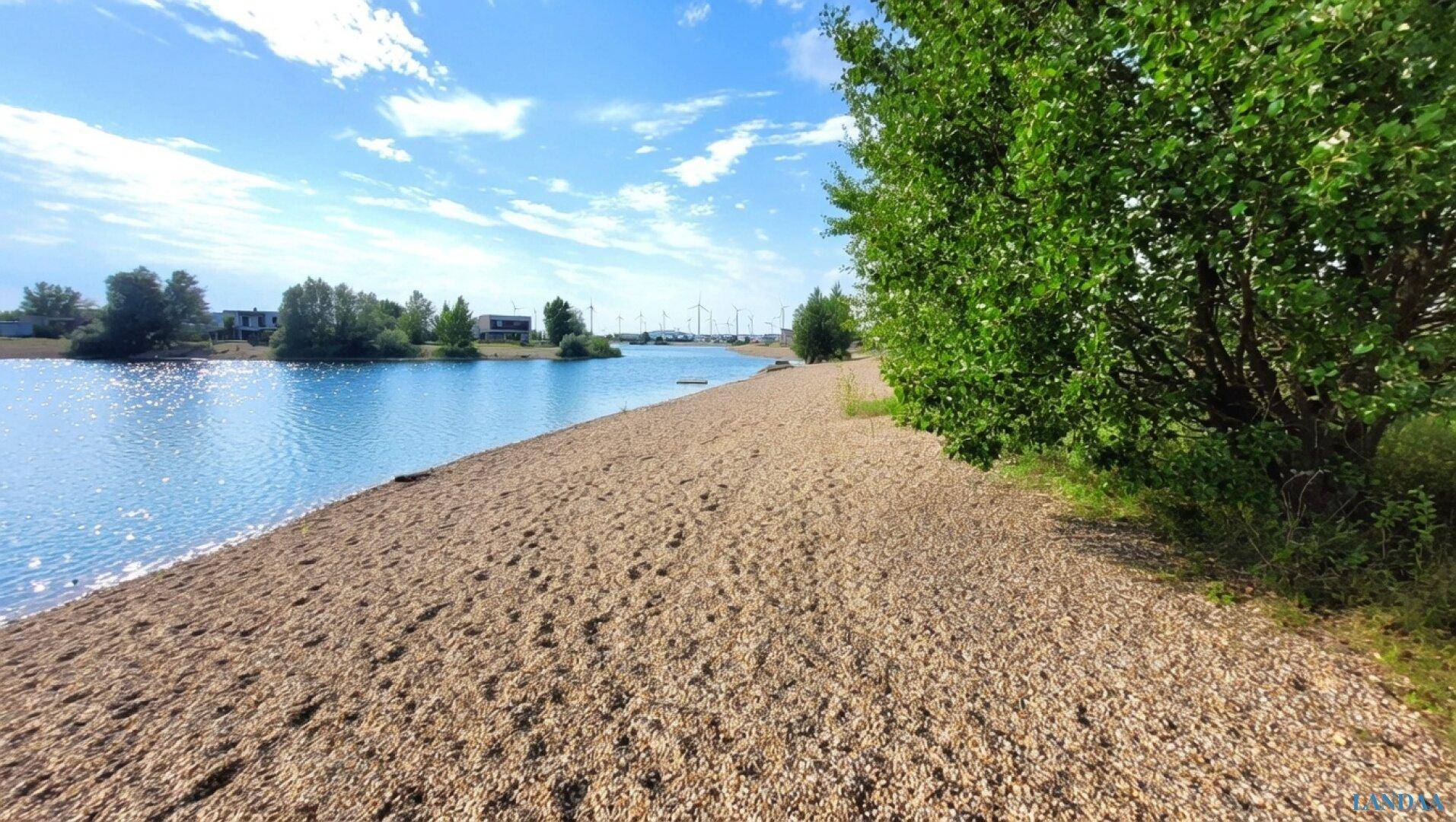 Langer Kiesstrand am Seeufer unter blauem Himmel mit vereinzelten Bäumen.