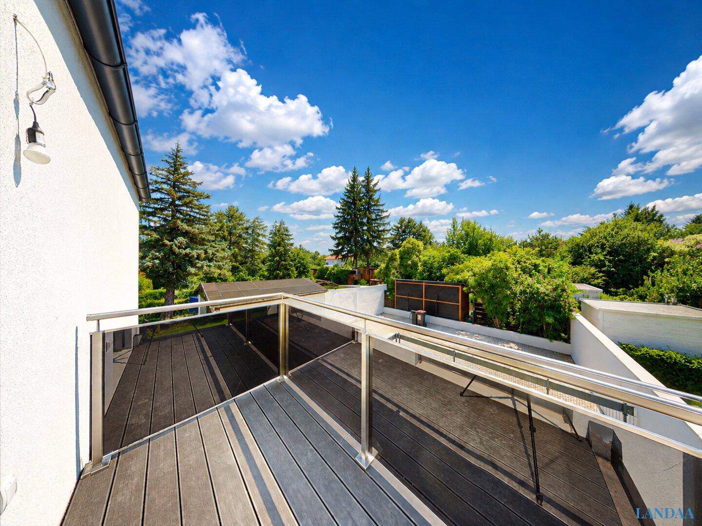 Geräumiger Balkon mit Holzboden und Glasgeländer, umgeben von grünen Bäumen.