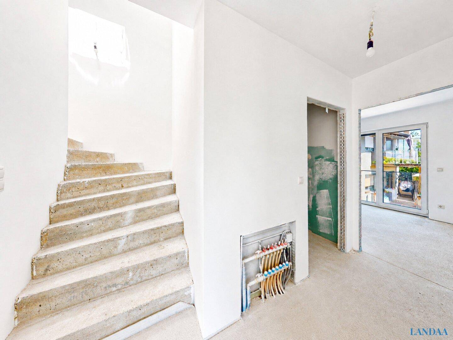 Treppenhaus mit Betontreppe und Blick auf Installationsleitungen in einem angrenzenden Raum.
