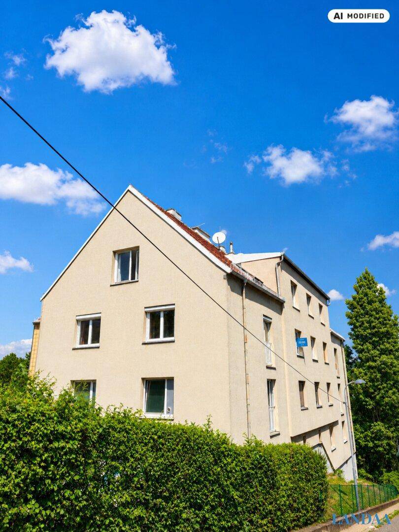 Seitenansicht eines Mehrfamilienhauses mit beiger Fassade, Satteldach und grüner Hecke.