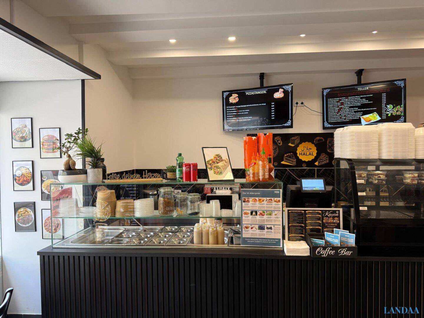 Innenansicht des Gastraums mit modernem Tresen, Speisevitrine und digitalen Menüanzeigen an der Wand.
