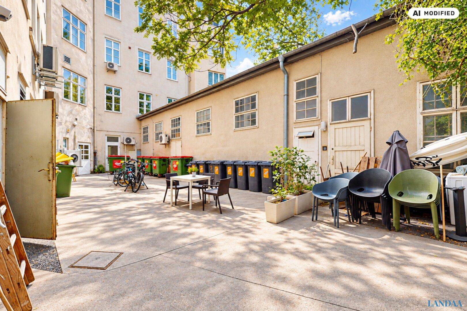 Hinterhof mit Mülltonnen, Fahrrädern und einem kleinen Sitzbereich, umgeben von Gebäuden und Bäumen.