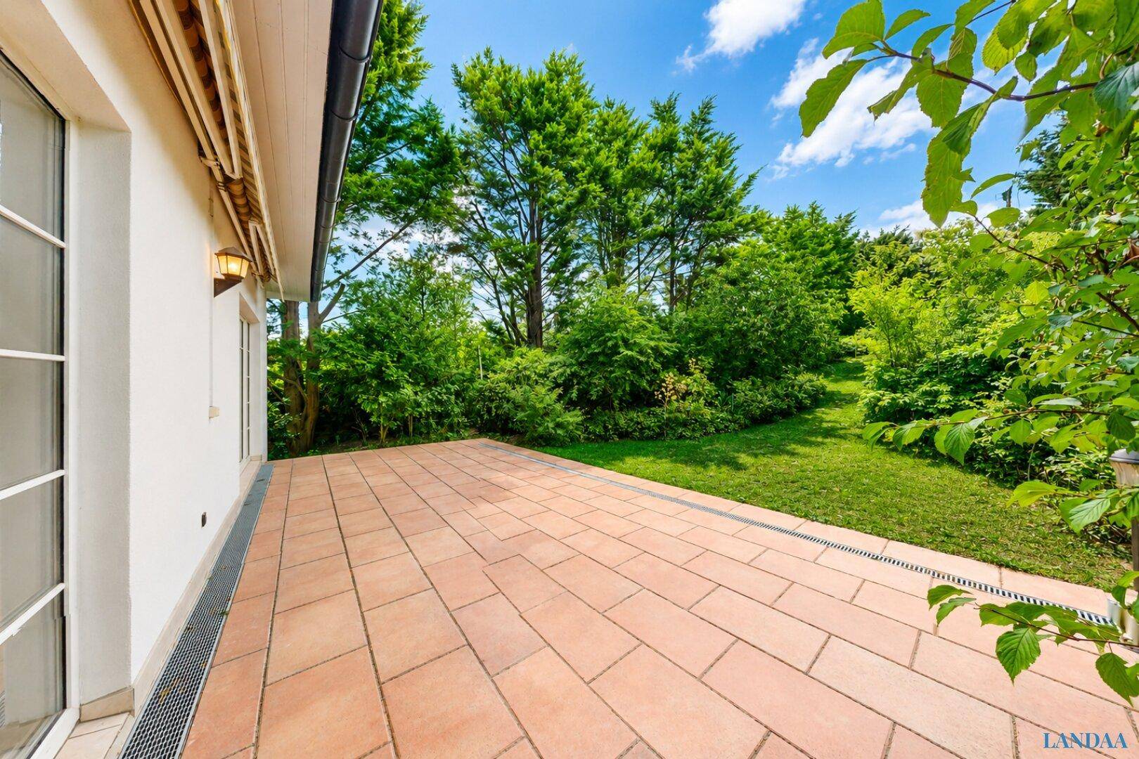 Geräumige, sonnige Terrasse mit hellbraunen Fliesen und direktem Übergang zum gepflegten Garten.