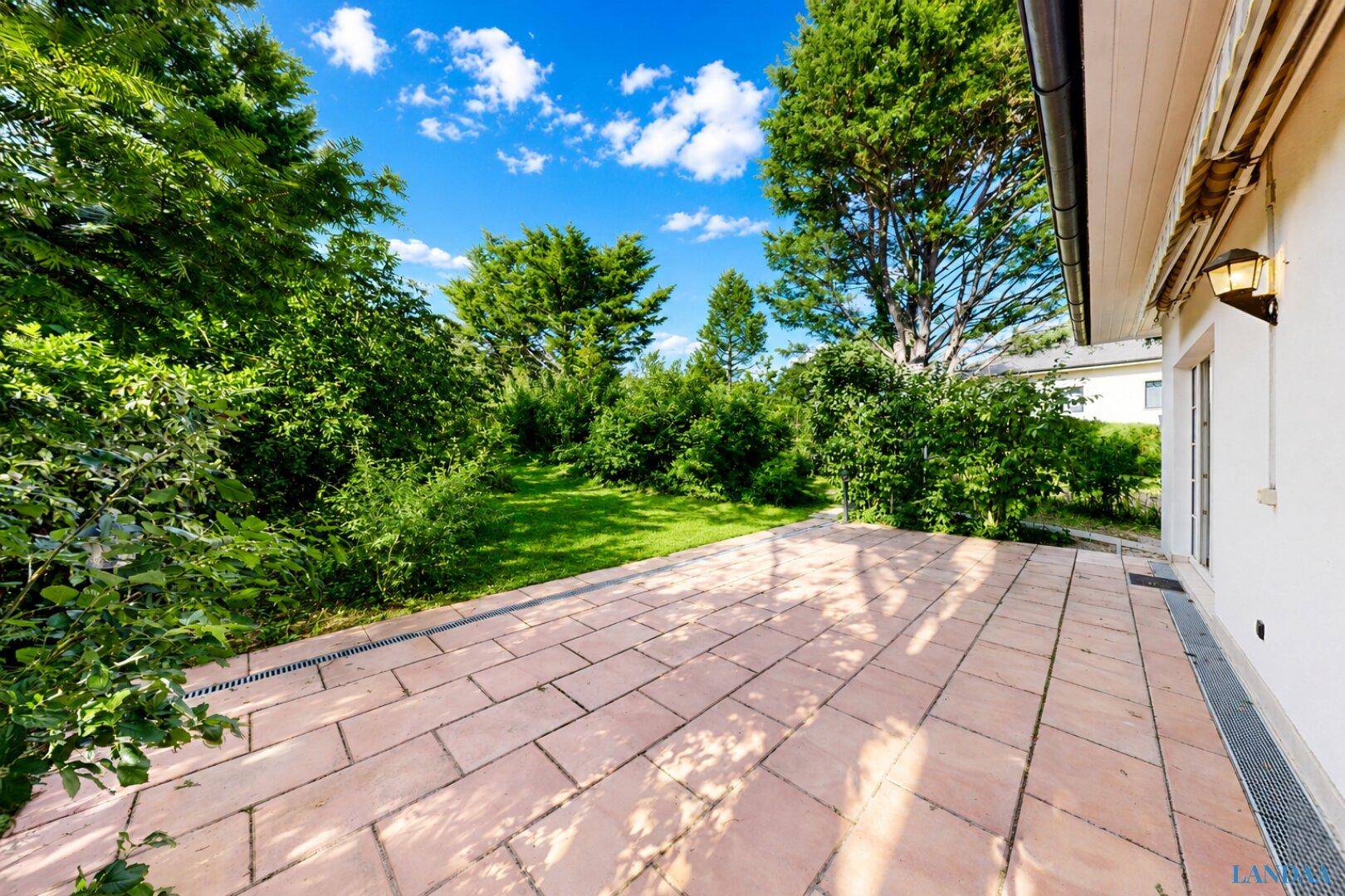 Weitläufige Terrasse mit hellen Fliesen, die in einen großen, grünen Garten übergeht.