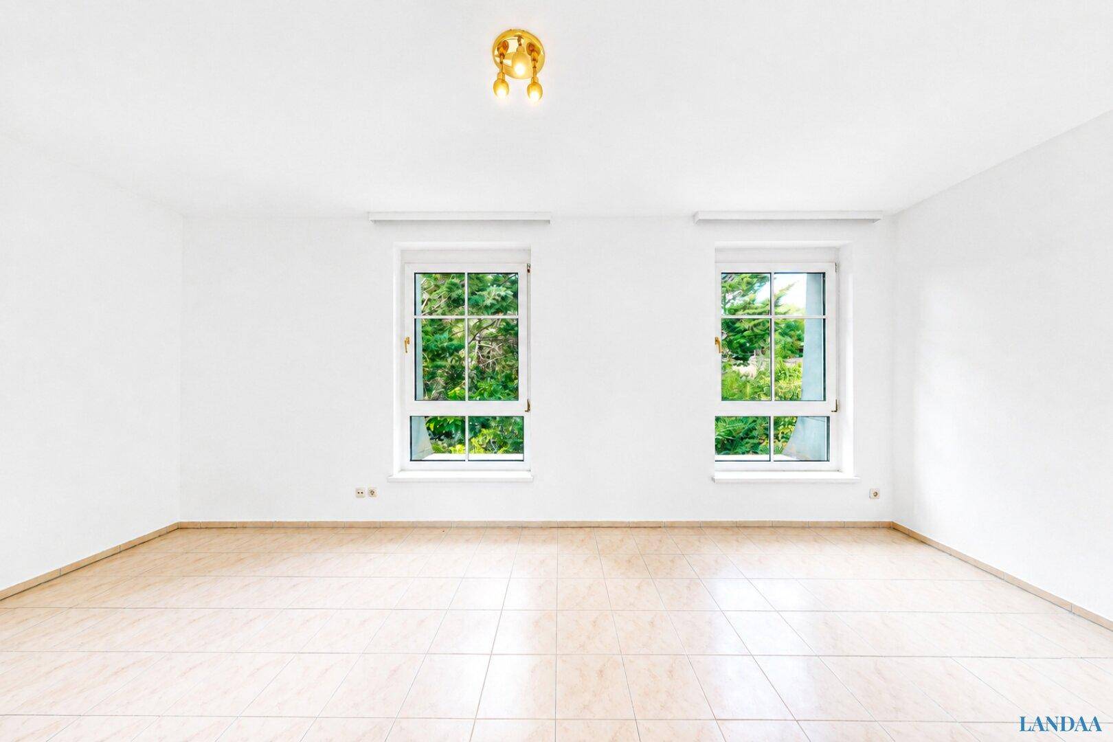 Helles, leeres Zimmer mit weißen Wänden, Fliesenboden und zwei Fenstern mit Blick ins Grüne.