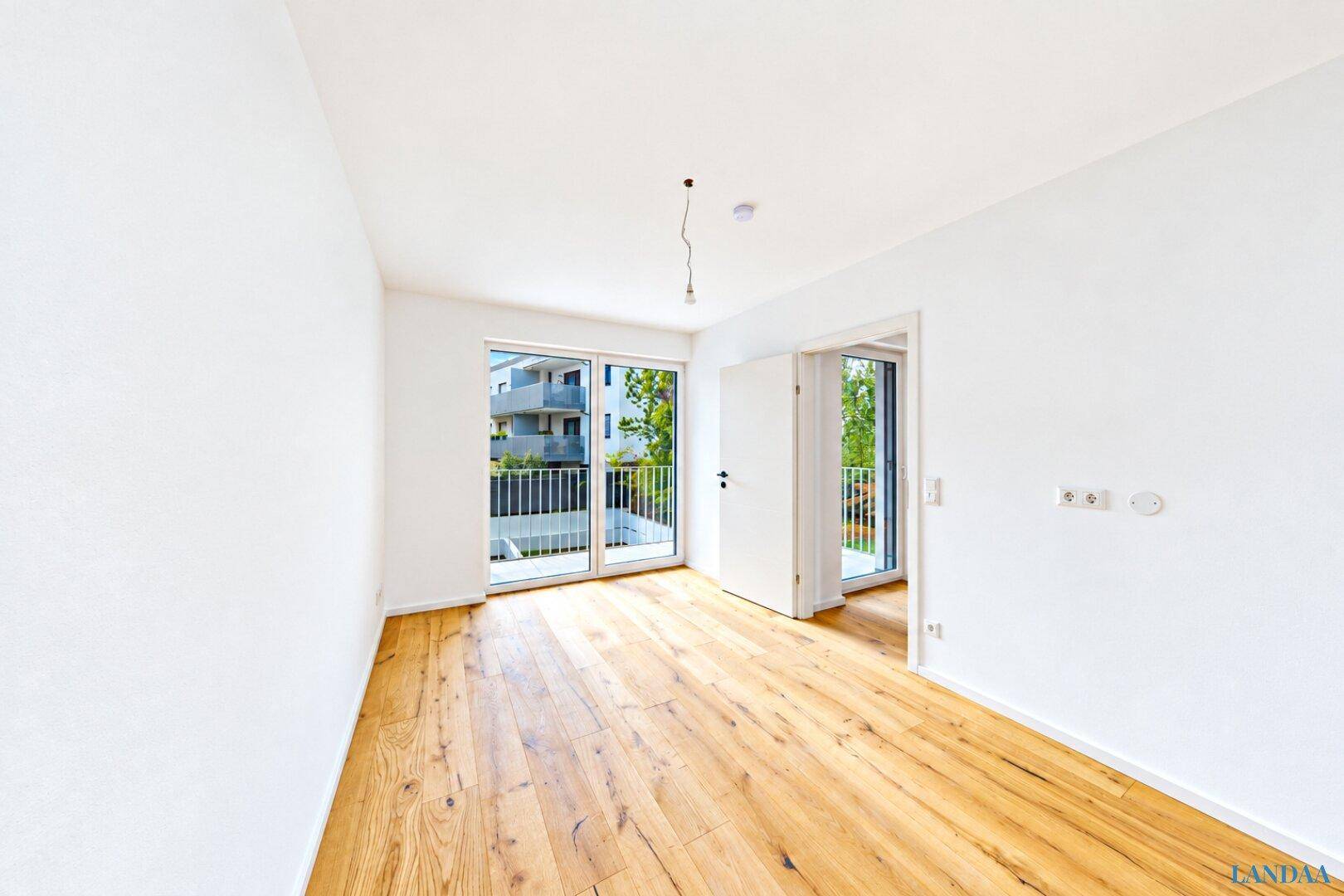 Leeres Schlafzimmer mit Holzboden und großen Fenstern, die Zugang zu einem Balkon bieten.