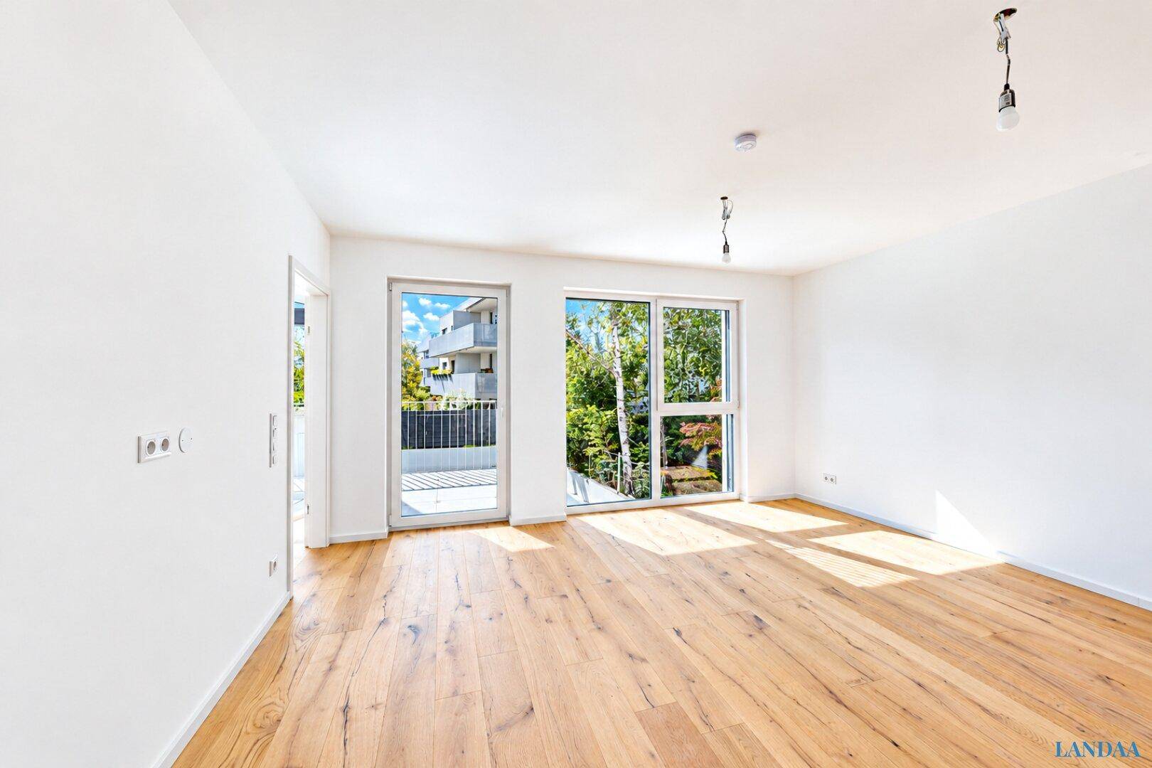 Geräumiger, leerer Wohn-Essbereich mit hellem Holzboden und großen Fenstern zum Balkon.