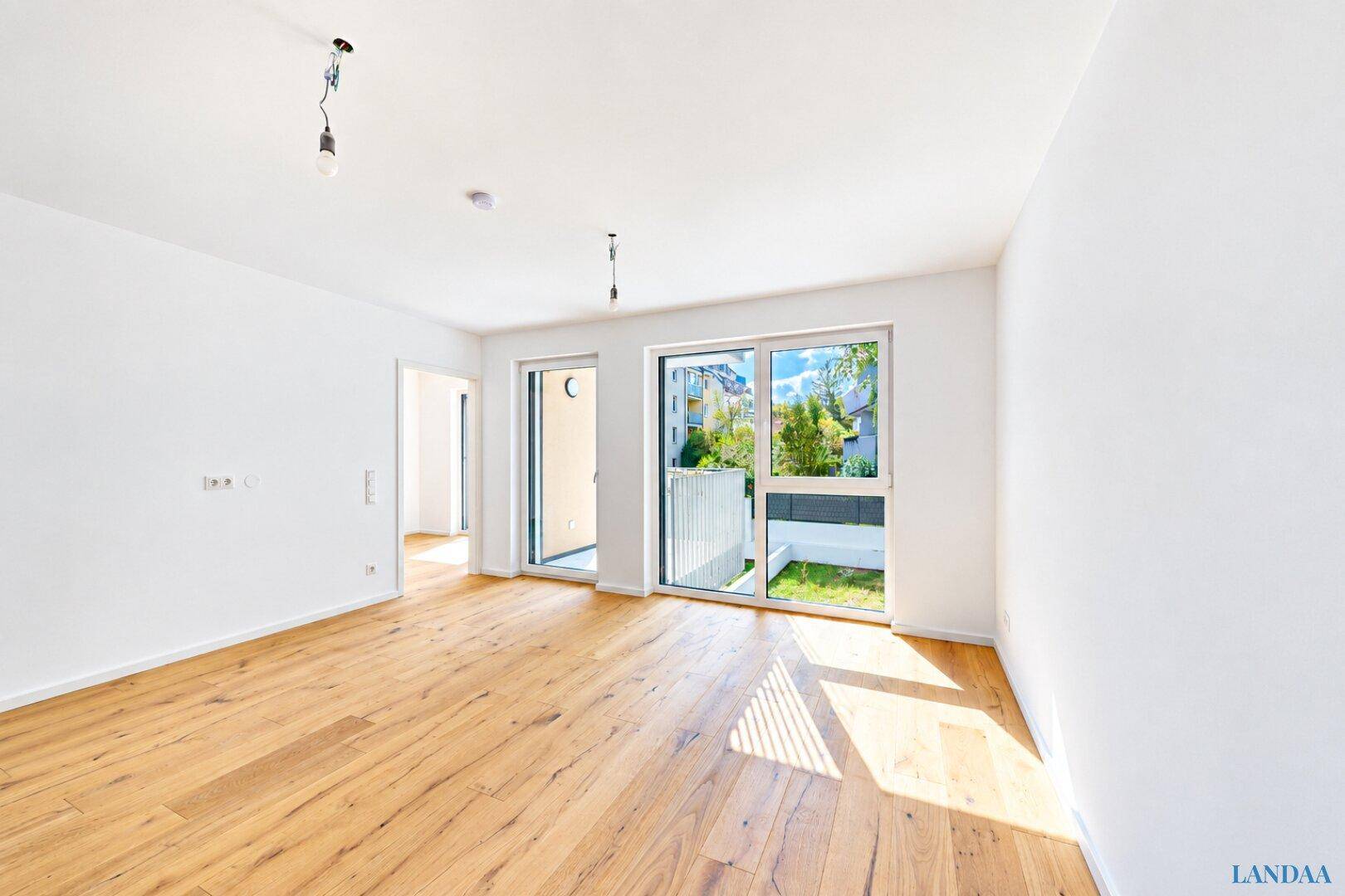 Geräumiger, leerer Raum mit Holzboden und Zugang zu einem kleinen privaten Gartenbereich.