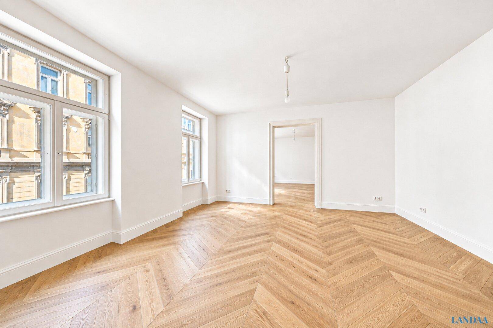Geräumiges Zimmer mit elegantem Fischgrätparkett und viel Tageslicht durch große Fenster.
