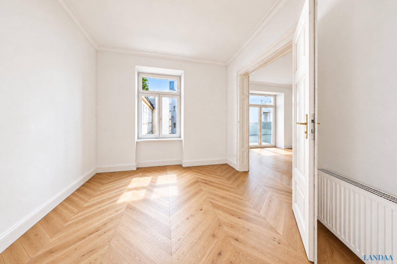 Helles Zimmer mit Fischgrätparkett und Zugang zu einem weiteren Raum mit Balkon.