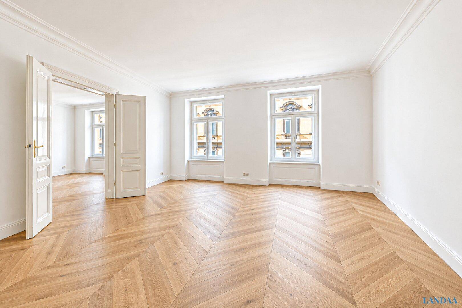 Großes, helles Zimmer mit Fischgrätparkett und Doppeltüren, die zu einem weiteren Wohnbereich führen.