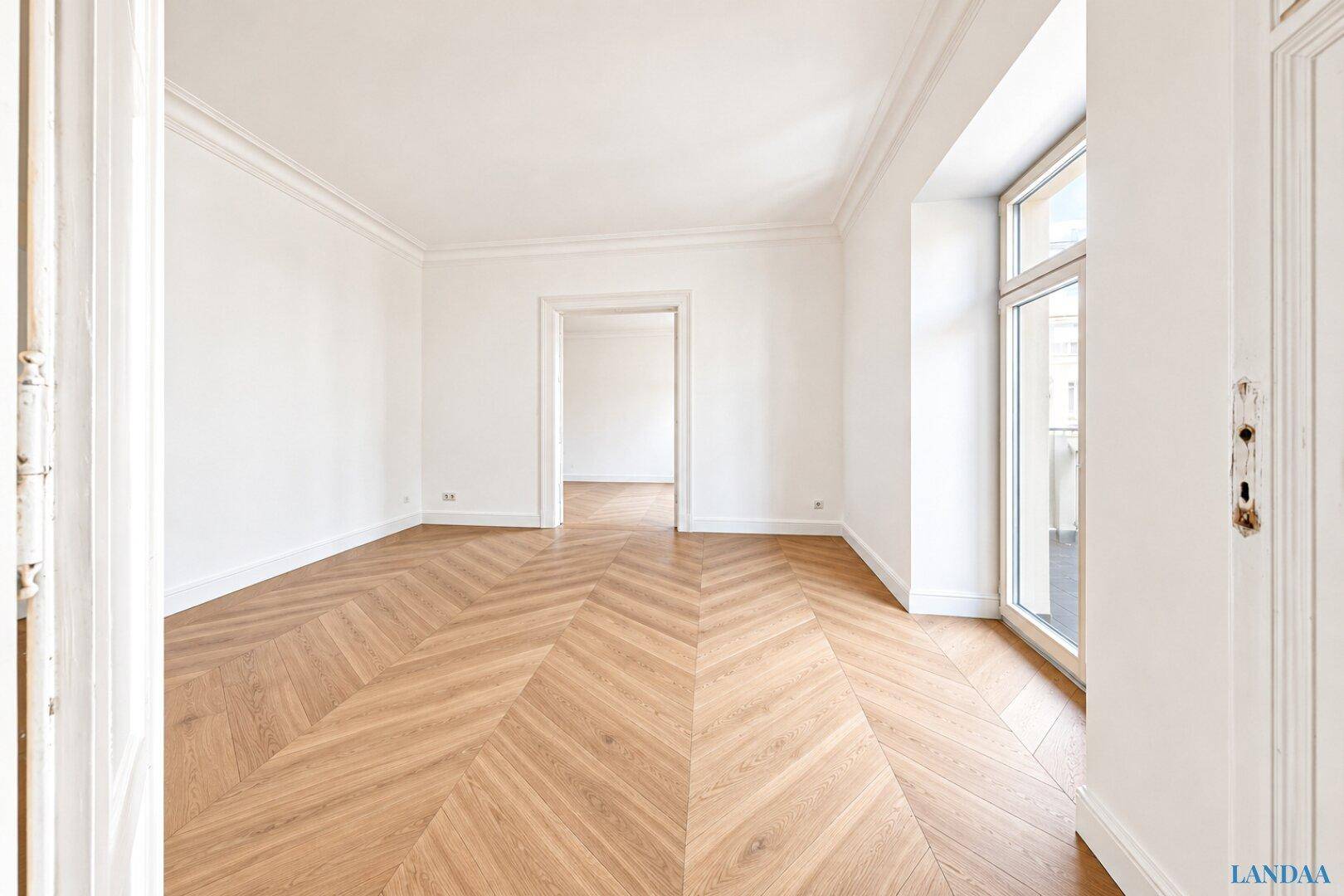 Geräumiges Zimmer mit Fischgrätparkett und einem großen Fenster, das zu einem französischen Balkon führt.