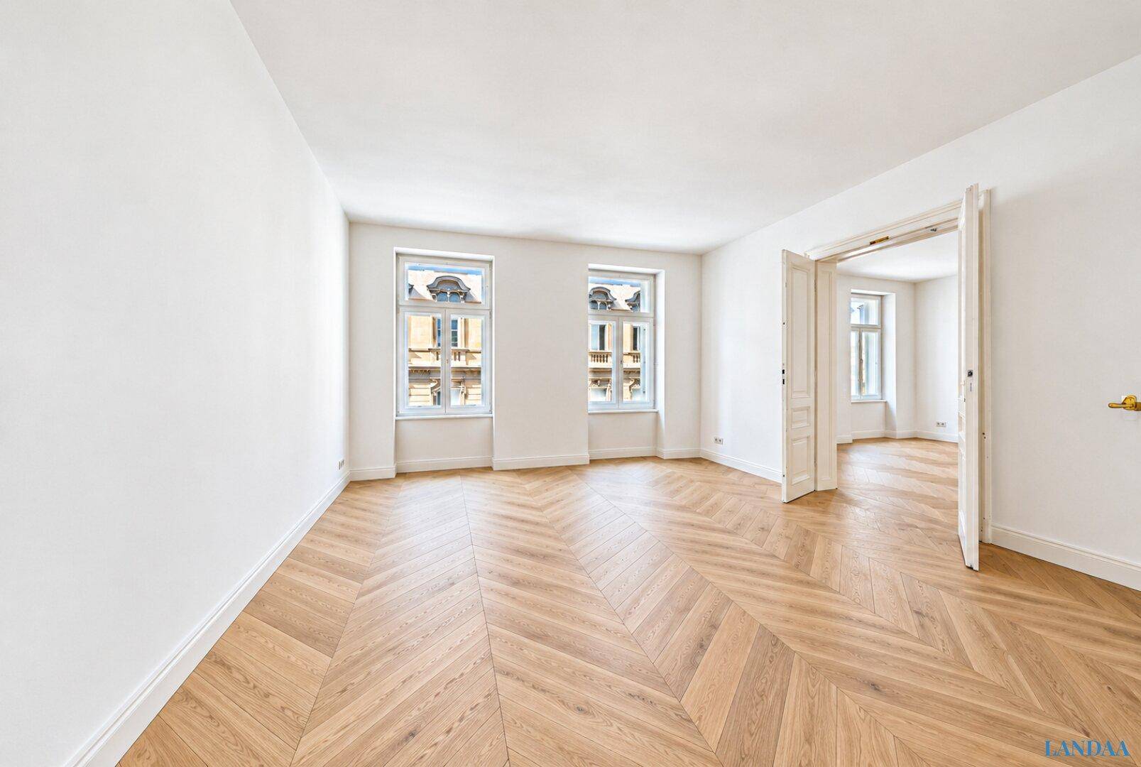 Helles Zimmer mit Fischgrätparkett, weißen Wänden und Doppeltüren zu einem angrenzenden Raum.