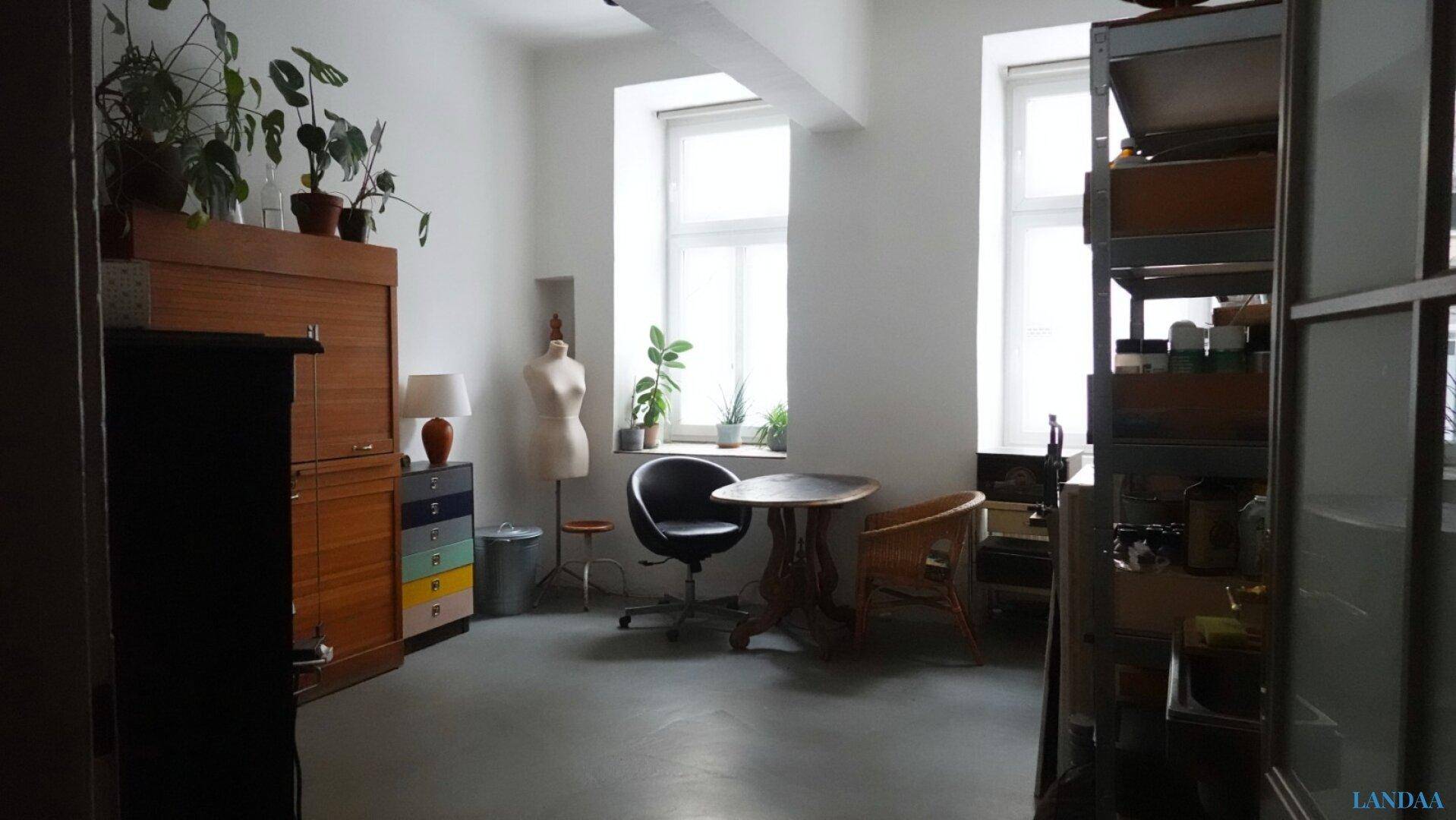 Heller Raum mit großen Fenstern, Pflanzen und einem Arbeitsbereich mit Schreibtisch und Stuhl.