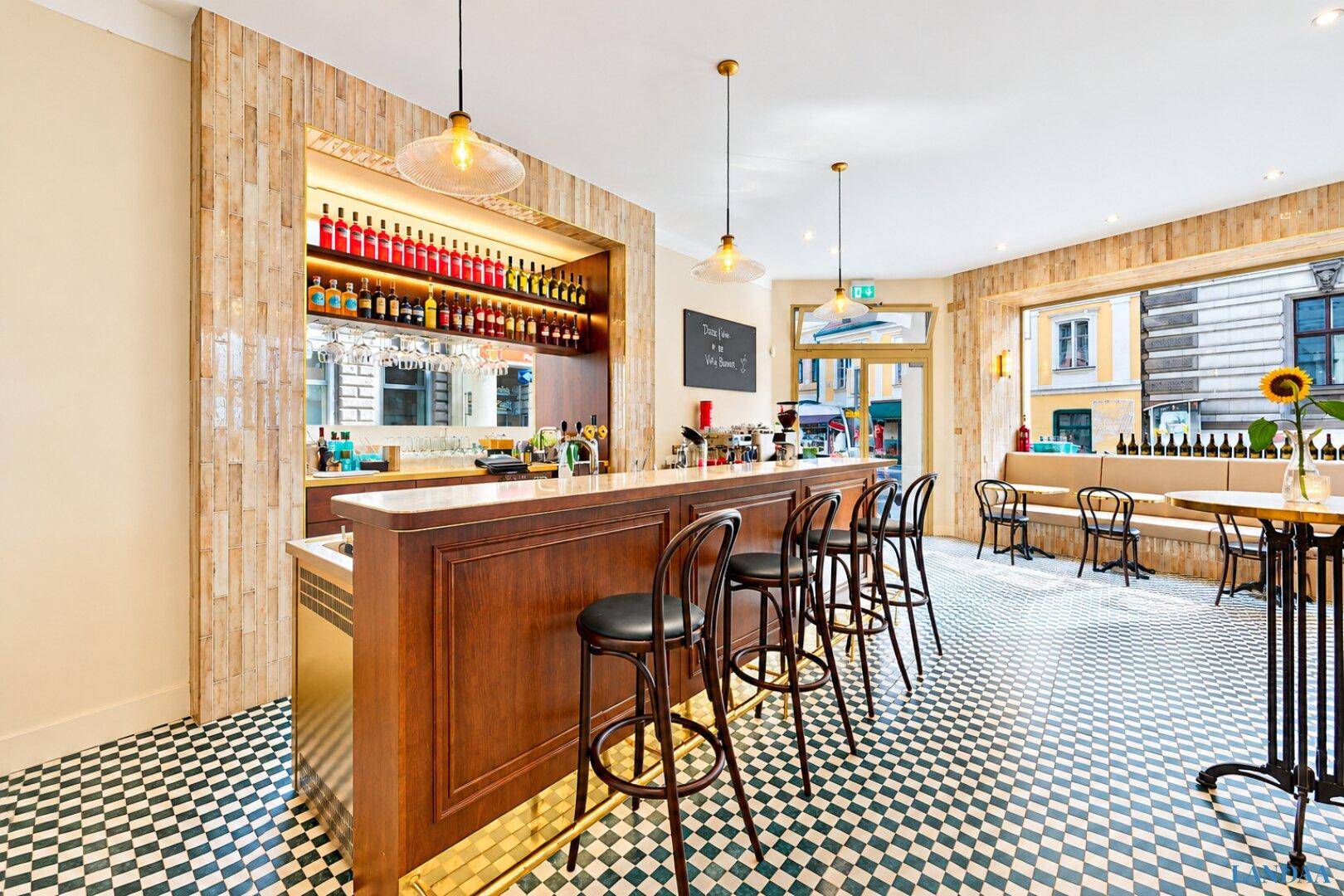 Moderne Bar mit Holztheke, hohen Barhockern und einem auffälligen Fliesenbodenmuster.