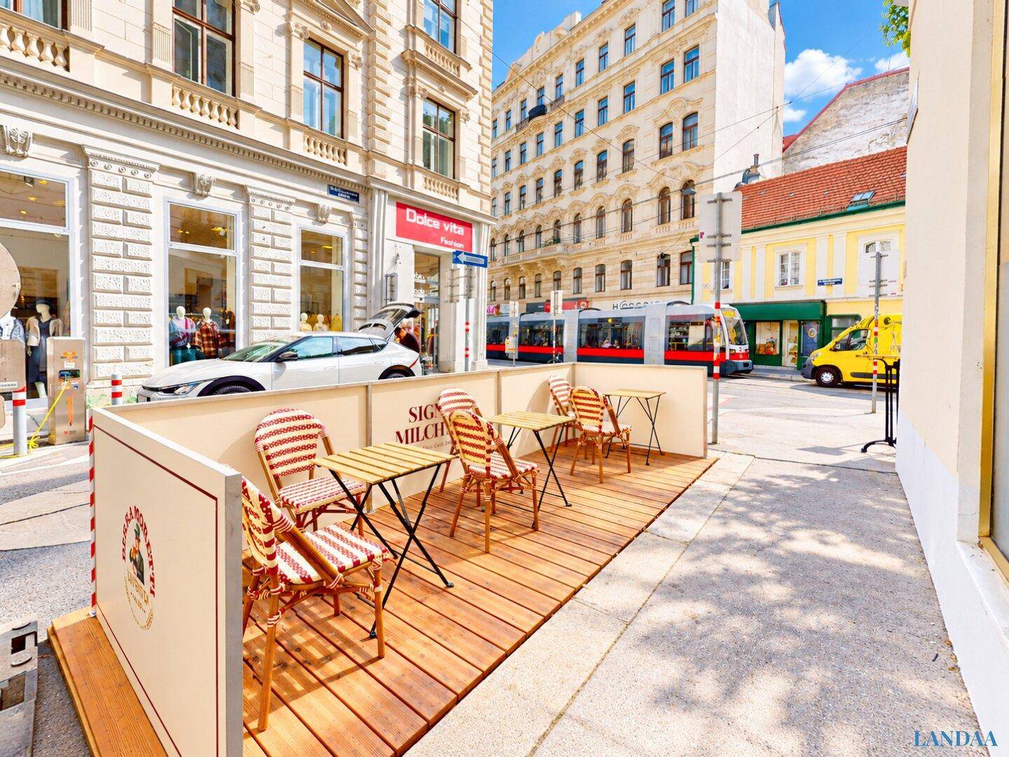 Außenbereich mit Holzboden und Tischen für Gäste, umgeben von einer belebten Stadtstraße.