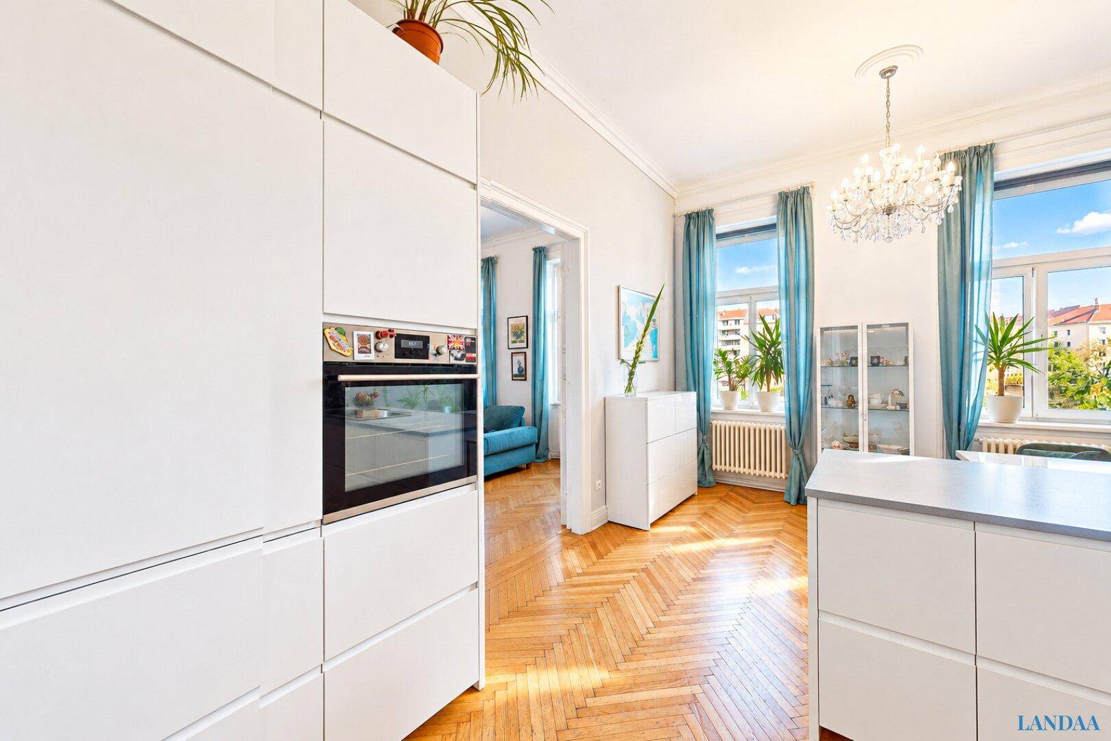 Moderne weiße Küche mit integriertem Backofen und Blick auf einen hellen Wohnbereich mit Parkettboden.