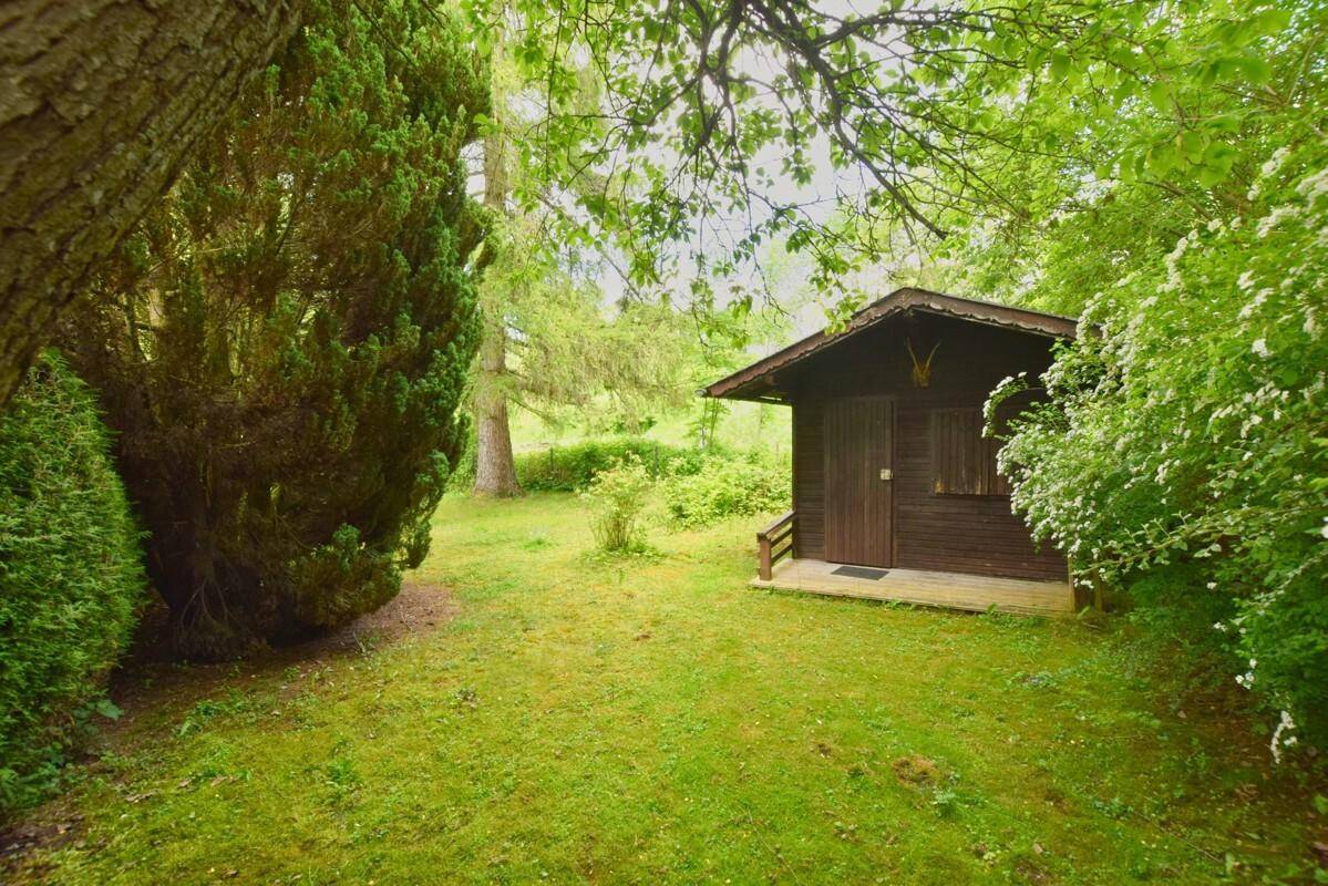 Kleines Holzhaus in einem grünen Garten, umgeben von Bäumen und Sträuchern.