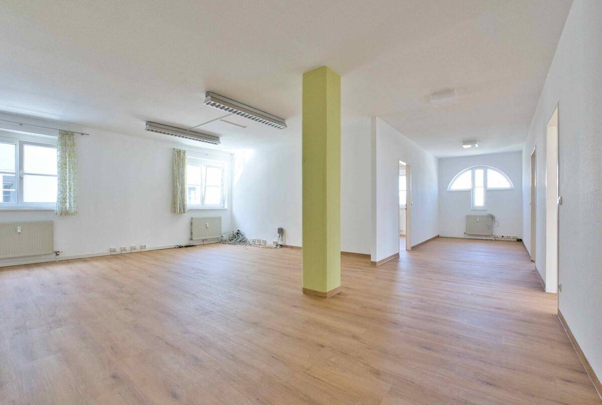 Heller Büroraum mit Laminatboden, weißen Wänden und verschiedenen Fenstern, inklusive eines Rundbogenfensters.