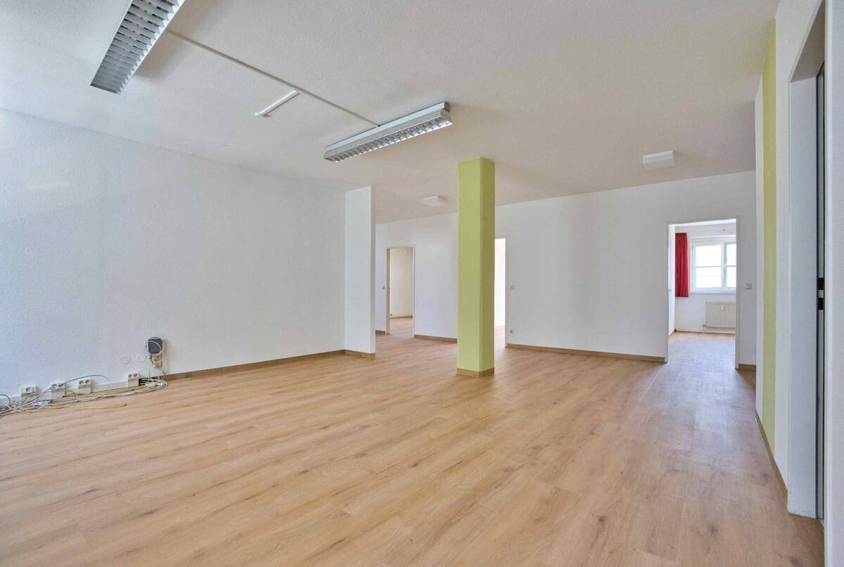 Großer, leerer Büroraum mit hellem Laminatboden, weißen Wänden und einem grünen Pfeiler.