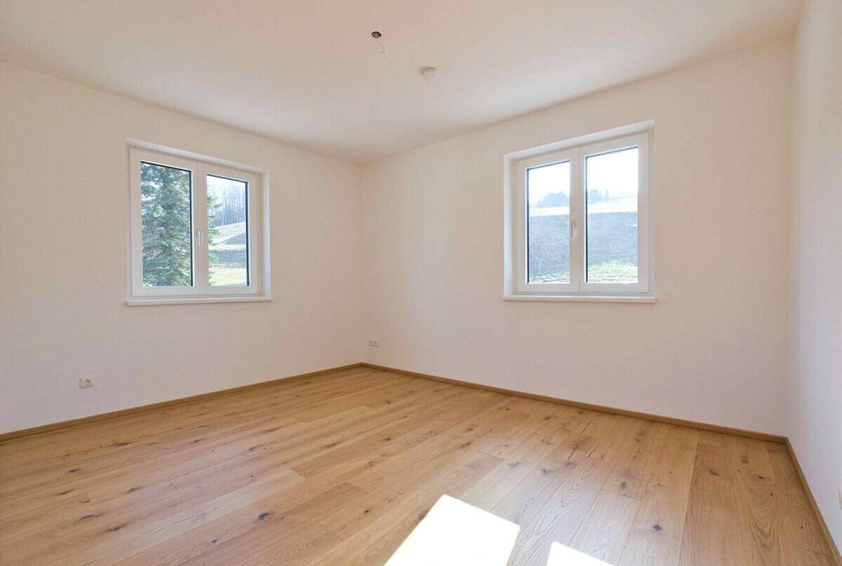 Helles Schlafzimmer mit zwei Fenstern, Holzboden und Blick ins Grüne, unmöbliert.