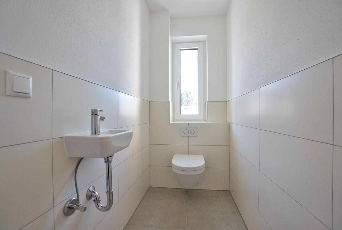 Modernes Gäste-WC mit wandhängendem WC, kleinem Waschbecken und Fenster, hell gefliest.