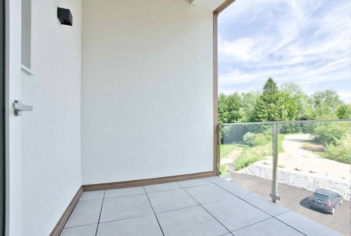 Moderner Balkon mit hellen Fliesen und elegantem Glasgeländer, umgeben von grüner Landschaft.