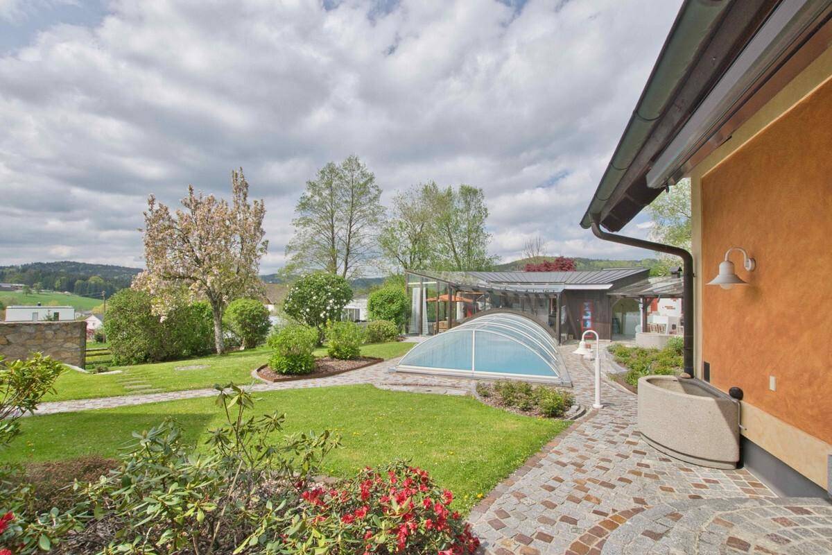 Gartenansicht mit überdachtem Pool, blühenden Sträuchern und weitem Blick in die Landschaft.