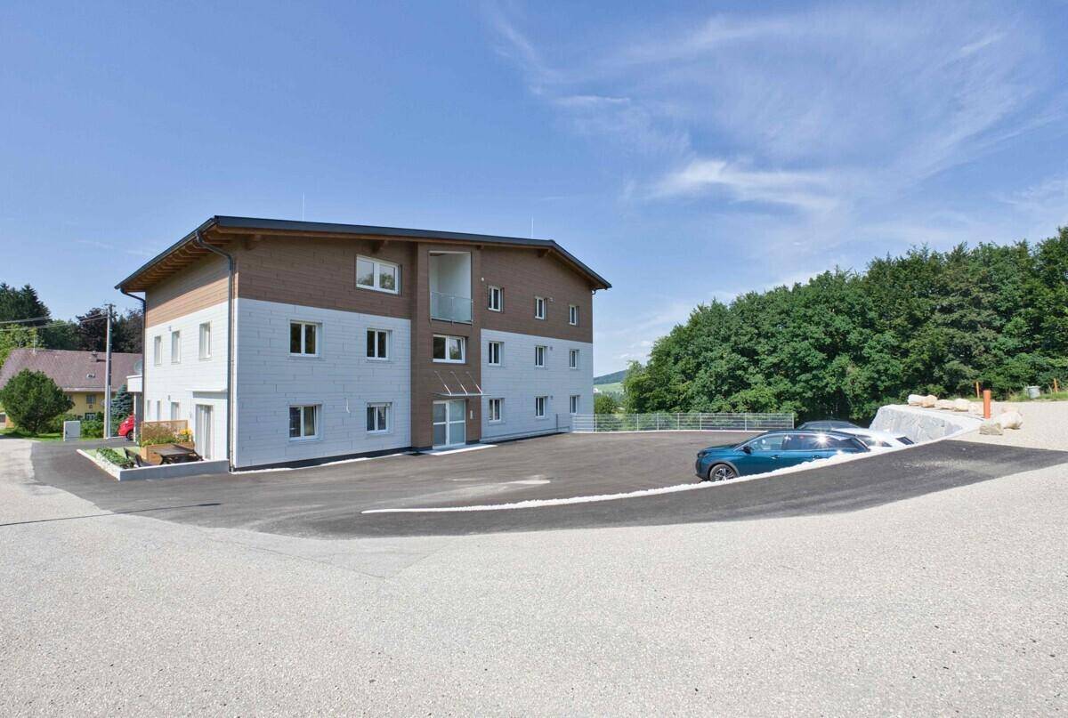 Modernes Mehrfamilienhaus mit Holzfassade und großem Parkplatz in grüner Umgebung.
