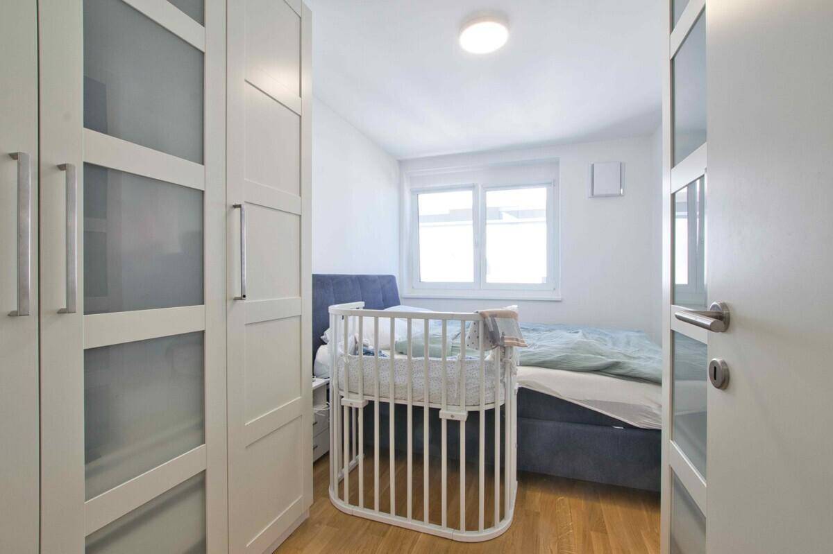 Schlafzimmer mit Doppelbett, weißem Kleiderschrank und einem Babybett am Fenster.