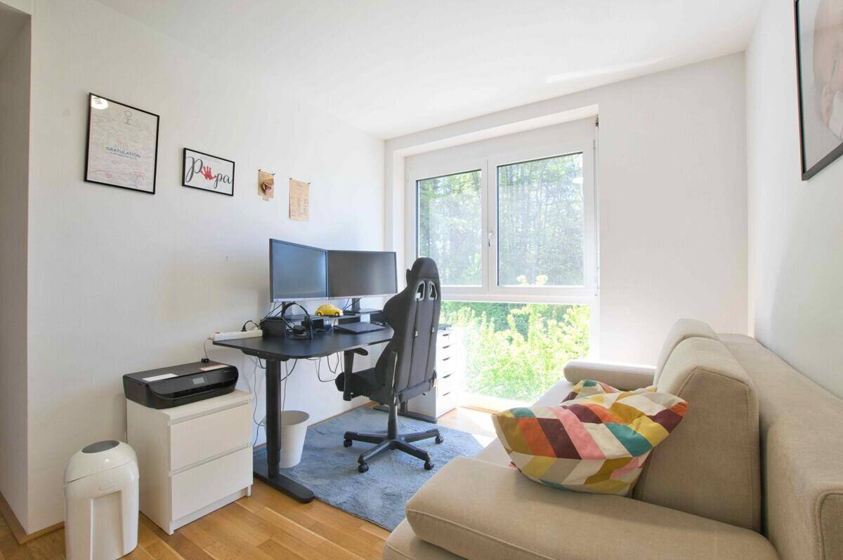 Modernes Arbeitszimmer mit Schreibtisch, zwei Monitoren und Sofa, bietet einen grünen Ausblick.