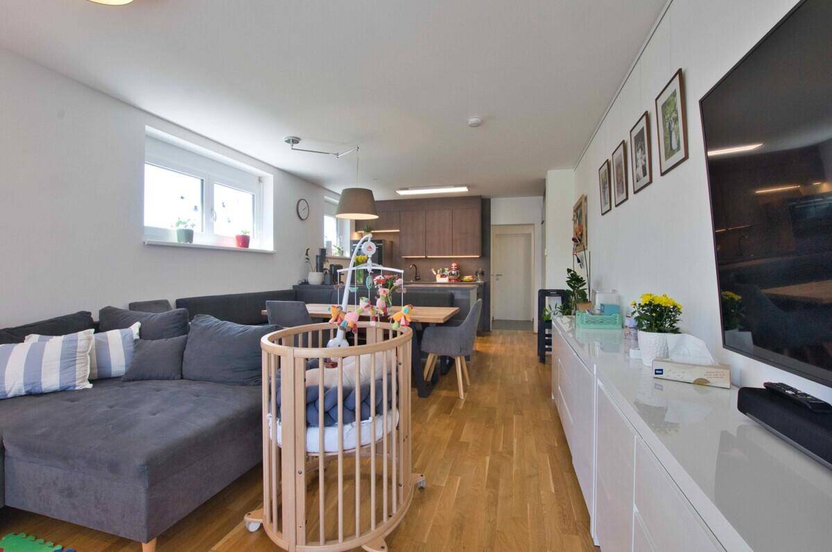 Heller Wohnbereich mit Sofa, Babybett und Sideboard, verbunden mit dem Küchenbereich.
